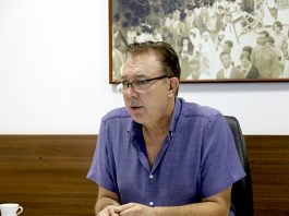 Orvino, sentado e debruçado com os cotovelos em sua mesa, usando camisa azul e óculos, fala durante entrevista