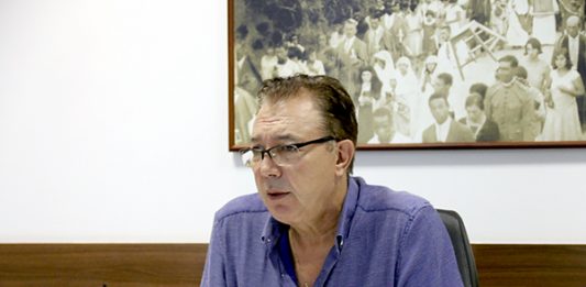 Orvino, sentado e debruçado com os cotovelos em sua mesa, usando camisa azul e óculos, fala durante entrevista