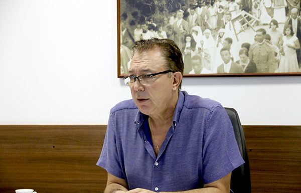 Orvino, sentado e debruçado com os cotovelos em sua mesa, usando camisa azul e óculos, fala durante entrevista