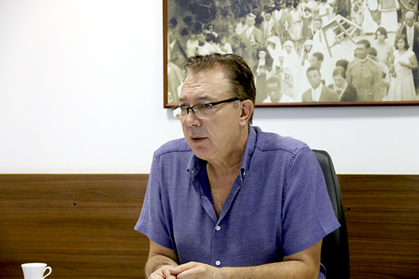 Orvino, sentado e debruçado com os cotovelos em sua mesa, usando camisa azul e óculos, fala durante entrevista