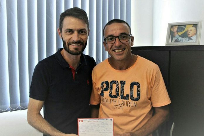 Dois homens posam para foto segurando a ficha de filiação de reni (à direita), usando camiseta laranja óculos; os dois estão no gabinete