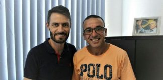 Dois homens posam para foto segurando a ficha de filiação de reni (à direita), usando camiseta laranja óculos; os dois estão no gabinete