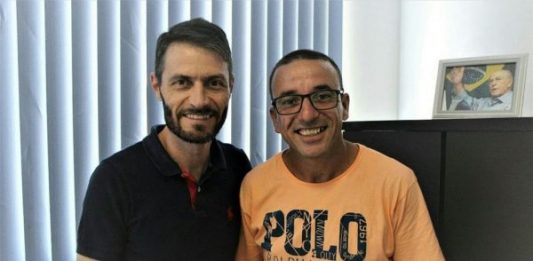 Dois homens posam para foto segurando a ficha de filiação de reni (à direita), usando camiseta laranja óculos; os dois estão no gabinete