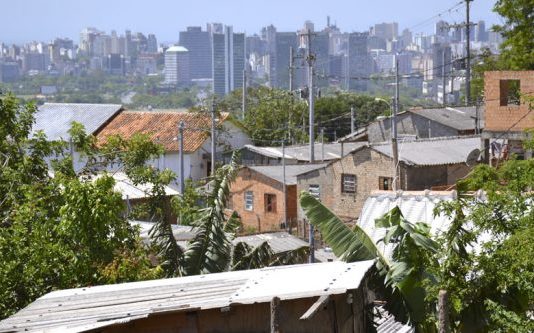 Vista da cidade, com casas pobres em primeiro plano e prédio de classe alta ao fundo