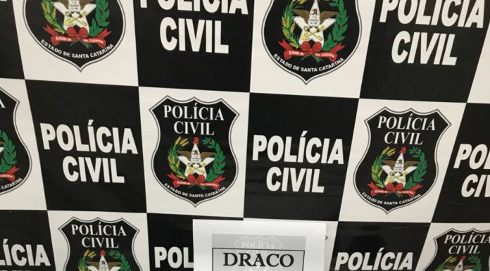 Mesa com armas, celulares e munições com um painel de logos da polícia civil ao fundo