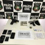 Mesa com armas, celulares e munições com um painel de logos da polícia civil ao fundo