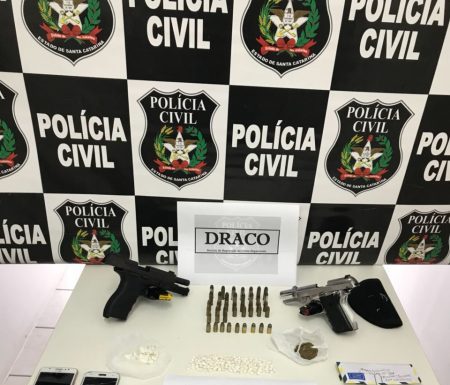 Mesa com armas, celulares e munições com um painel de logos da polícia civil ao fundo