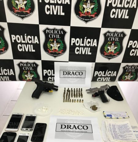 Mesa com armas, celulares e munições com um painel de logos da polícia civil ao fundo