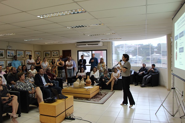 Programa Cidade Empreendedora São José Sebrae-SC Aemflo CDL-SJ Mulher faz apresentação para uma plateia, parte sentada, parte em pé, vista de lado segurando um microfone e à frente de um telão