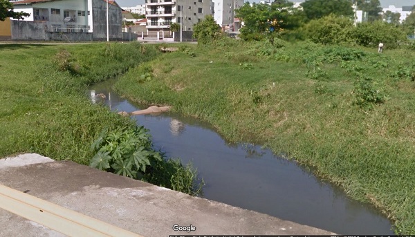 Curso de água do Rio Buchler poluído, com cano atravessando o pequeno leito envolto em mato num terreno baldio