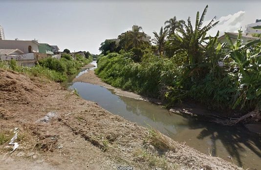 Bacia do Rio Buchler terá programa de regularização de esgoto Curso de água do Rio Buchler poluído, quase seco, com algum mato e casas em volta