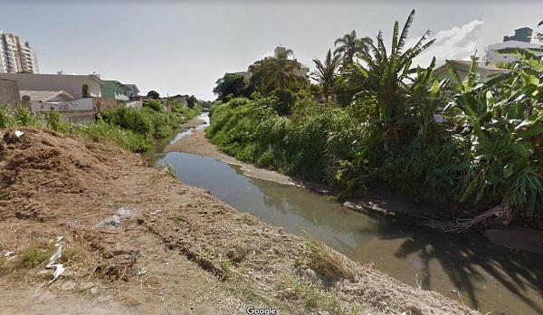 Curso de água do Rio Buchler poluído, quase seco, com algum mato e casas em volta