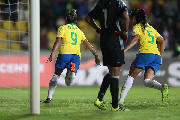 Duas jogadoras da seleção brasileira saem correndo comemorando gol, com goleira adversária em primeiro plano