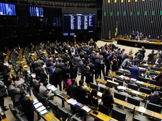 Vista do salão do Senado, com muitas cadeiras e pessoas engravatadas em pé pelo ambiente