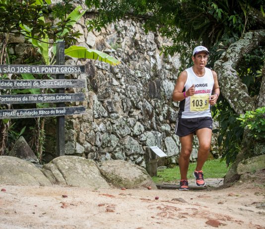 Atleta corre em uma trilha de florianópolis, com um muro de pedra ao fundo e placas de direções ao lado