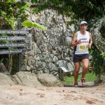 Atleta corre em uma trilha de florianópolis, com um muro de pedra ao fundo e placas de direções ao lado