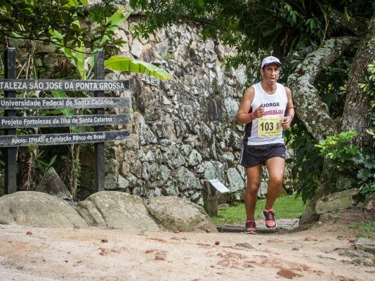 Atleta corre em uma trilha de florianópolis, com um muro de pedra ao fundo e placas de direções ao lado