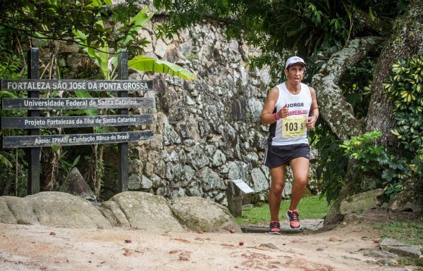 Atleta corre em uma trilha de florianópolis, com um muro de pedra ao fundo e placas de direções ao lado