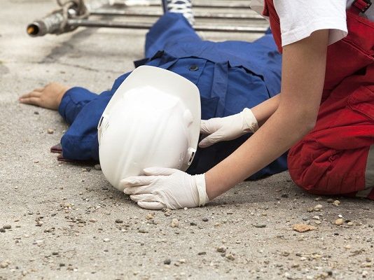 A cada três dias um trabalhador morre em Santa Catarina Operário de construção civil usando macacão azul e capacete branco estendido no chão sendo atendido por uma mulher de macacão vermelho e luvas brancas
