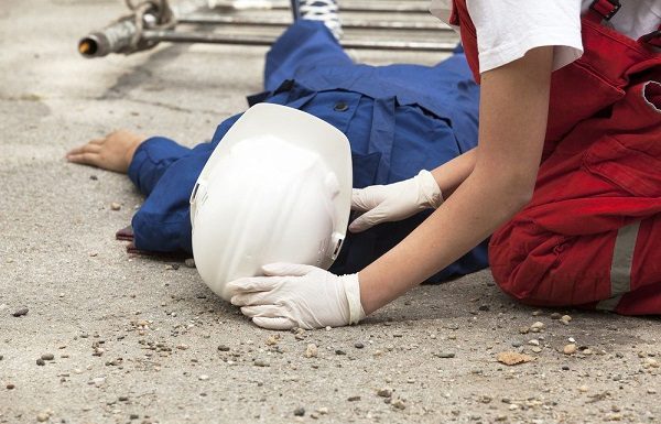 Operário de construção civil usando macacão azul e capacete branco estendido no chão sendo atendido por uma mulher de macacão vermelho e luvas brancas