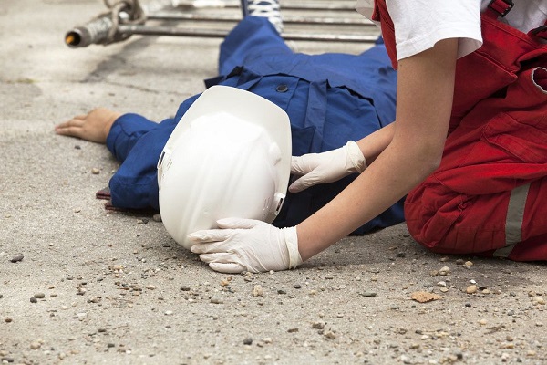 Operário de construção civil usando macacão azul e capacete branco estendido no chão sendo atendido por uma mulher de macacão vermelho e luvas brancas