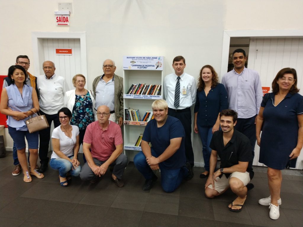 Grupo de pessoas em pé ou agachadas, entre homens e mulheres de diferentes idades, posa para foto ao lado da estante de livros