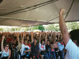 Servidores sentados em assembleia em que cada um está com o braço levantado e punho cerrado