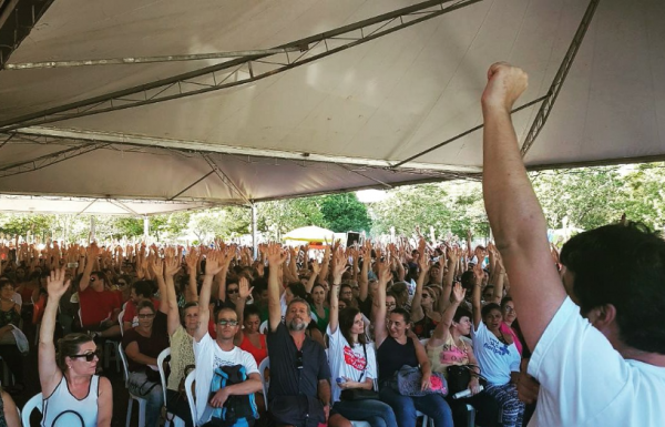 Servidores sentados em assembleia em que cada um está com o braço levantado e punho cerrado