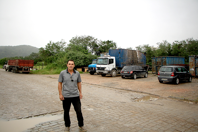 homem parado no meio da rua posa para foto, onde ao fundo há um terreno baldio com algumas árvores e dois caminhões e carros estacionados