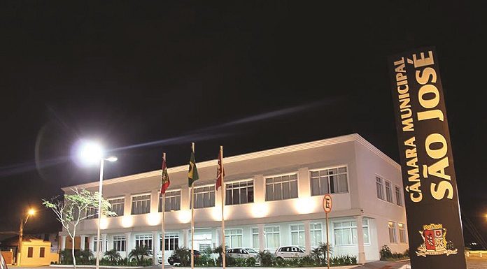 fachada do prédio da câmara vista de lado à noite, bem iluminada, com uma placa à direita escrita Câmara de Vereadores de São José, na vertical