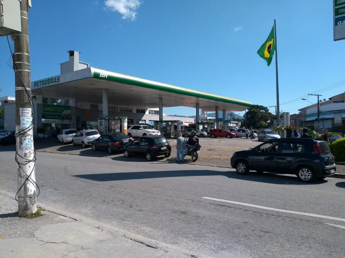 fila de carros em frente a um posto da petrobras com uma bandeira do brasil ao fundo