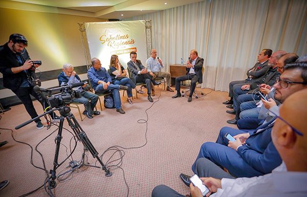 No centro, Merisio sentado em uma cadeira, e de cada lado 6 jornalistas sendados, à frente de uma câmera