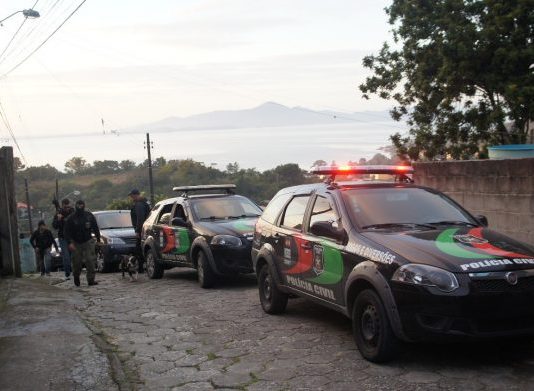 Operação na Fazenda Santo Antônio apreende documentação de facção criminosa ladeira estreita de paralelepípedos com carros da polícia civil estacionados e agentes usando balaclava e armamento pesado
