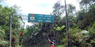 homem visto de costas em uma bicicleta com um braço levantado passa por uma estrada de chão em meio a um matagal e uma placa que diz "volte sempre"