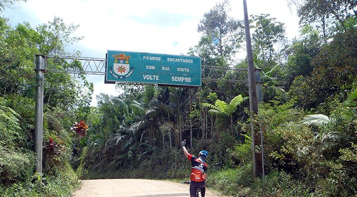 homem visto de costas em uma bicicleta com um braço levantado passa por uma estrada de chão em meio a um matagal e uma placa que diz "volte sempre"