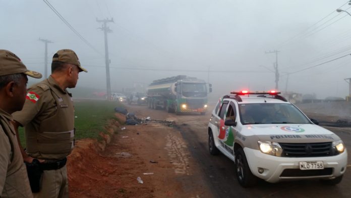 carro da polícia escoltando caminhão tanque em um dia de neblina com polícias ao lado da estrada