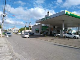 vista central de uma rua, com o posto de combustível de um lado e fila de carros do outro