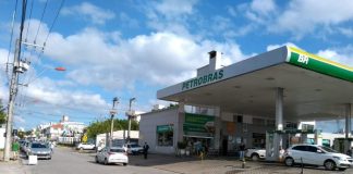 vista central de uma rua, com o posto de combustível de um lado e fila de carros do outro