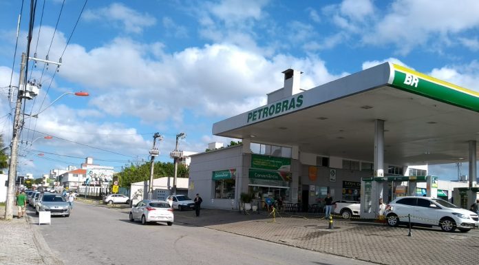 vista central de uma rua, com o posto de combustível de um lado e fila de carros do outro