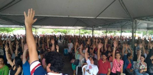 centenas de pessoas sentadas em assembleia com o braço estendido para o alto; em primeiro plano, um homem de costas segurando um microfone com a mão para o alto
