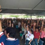 centenas de pessoas sentadas em assembleia com o braço estendido para o alto; em primeiro plano, um homem de costas segurando um microfone com a mão para o alto