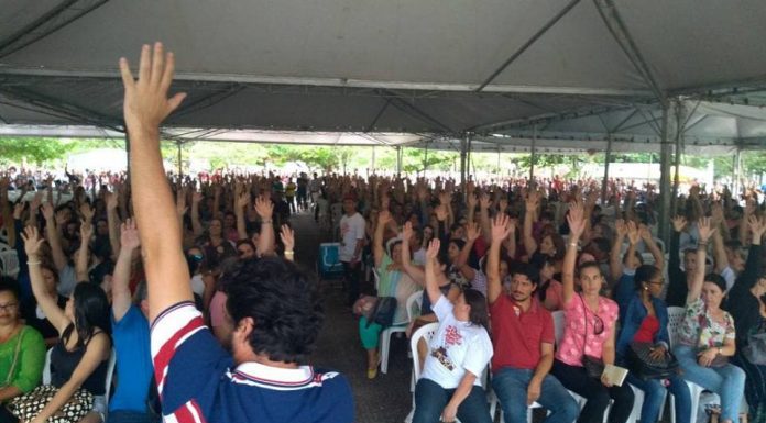 centenas de pessoas sentadas em assembleia com o braço estendido para o alto; em primeiro plano, um homem de costas segurando um microfone com a mão para o alto