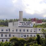 Vista externa e distante da fachada e parte superior do hospital, onde se lê o nome na parte mais alta do prédio, com árvores na frente e atrás