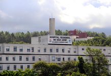 Vista externa e distante da fachada e parte superior do hospital, onde se lê o nome na parte mais alta do prédio, com árvores na frente e atrás