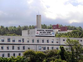 Vista externa e distante da fachada e parte superior do hospital, onde se lê o nome na parte mais alta do prédio, com árvores na frente e atrás