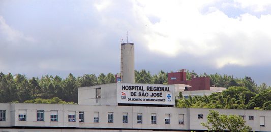Vista externa e distante da fachada e parte superior do hospital, onde se lê o nome na parte mais alta do prédio, com árvores na frente e atrás