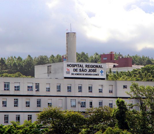 Vista externa e distante da fachada e parte superior do hospital, onde se lê o nome na parte mais alta do prédio, com árvores na frente e atrás