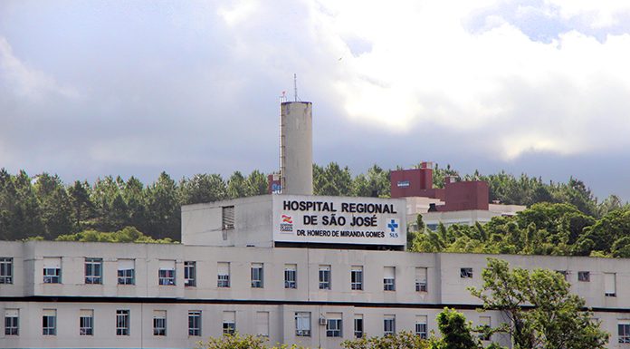 Vista externa e distante da fachada e parte superior do hospital, onde se lê o nome na parte mais alta do prédio, com árvores na frente e atrás