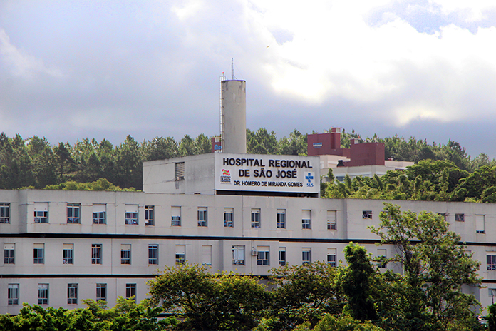 Vista externa e distante da fachada e parte superior do hospital, onde se lê o nome na parte mais alta do prédio, com árvores na frente e atrás