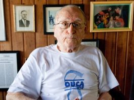 a imagem mostra o ex-governador Antônio Carlos Konder Reis sentado. Atrás dele estão fotografias penduradas na parede de madeira